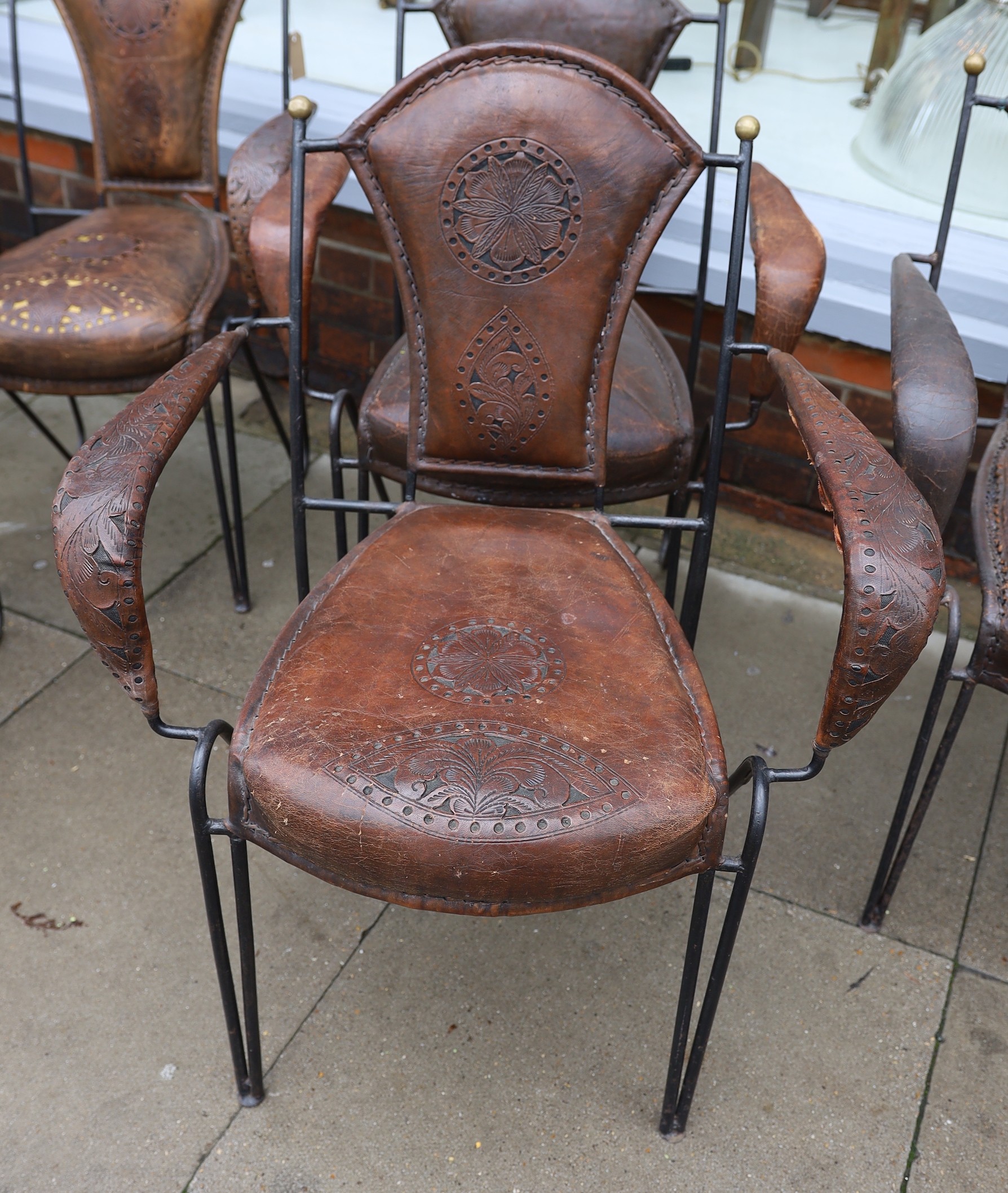 A set of six French wrought iron and brown leather elbow chairs, with brass ball finials, four with tooled and pierced decoration the others plain, height 84cm. width 61cm